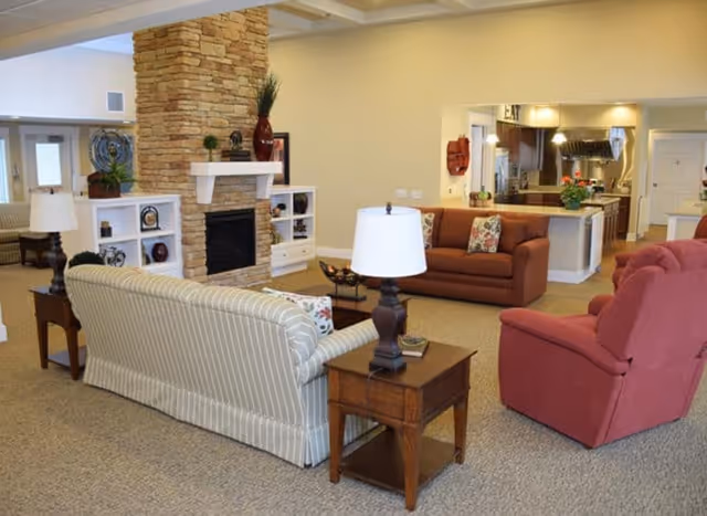 A cozy senior living common area featuring a stone fireplace with white built-in shelves on either side, a striped sofa, a red armchair, a brown sofa, wooden side tables with lamps, and a view into a kitchen area with hanging lights and a countertop with plants.