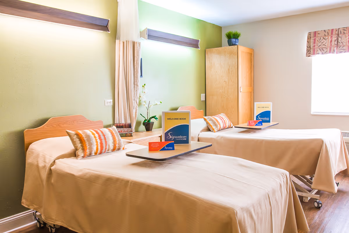 A bright and clean room with two single beds, each with a beige bedspread and a decorative pillow. Each bed has a small overbed table holding a welcome book and a card from Signature HealthCARE. The room has a green accent wall with a wooden headboard and a window with patterned curtains letting in natural light. There is a wooden cabinet and a small plant on top of it in the corner.