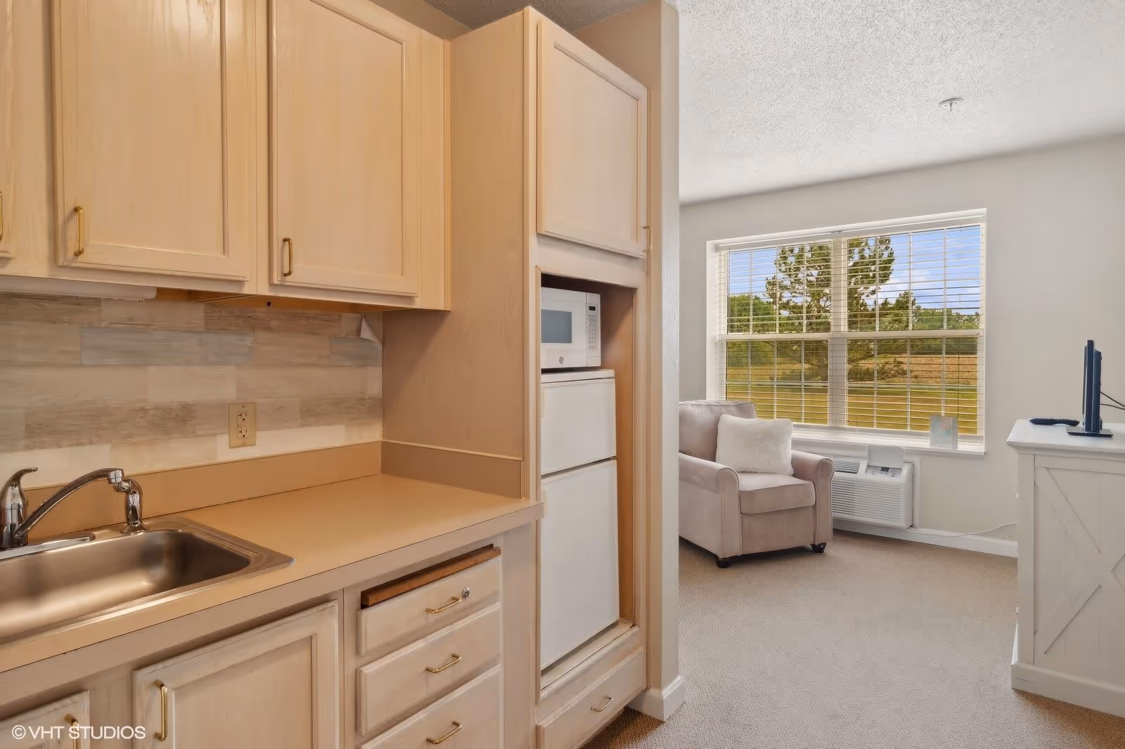 Interior view of a small kitchen area with light wood cabinets, a stainless steel sink, a microwave, and a mini refrigerator. Adjacent to the kitchen is a cozy living space with a beige armchair, a white pillow, a window with blinds showing an outdoor view of trees and grass, and a white TV stand with a television.