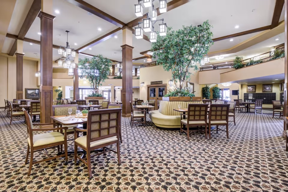 Spacious dining room with multiple tables and chairs, a central circular banquette, tall indoor trees, and decorative hanging lights.
