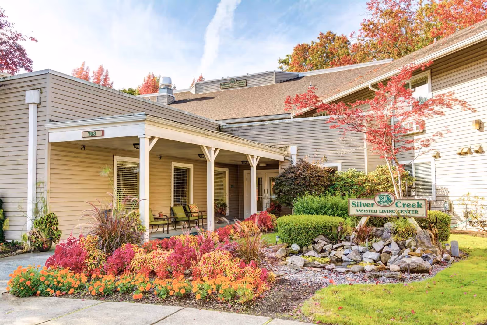 Front entrance of Silver Creek Assisted Living Center with a covered porch, seating, colorful landscaped flowerbeds and a sign.