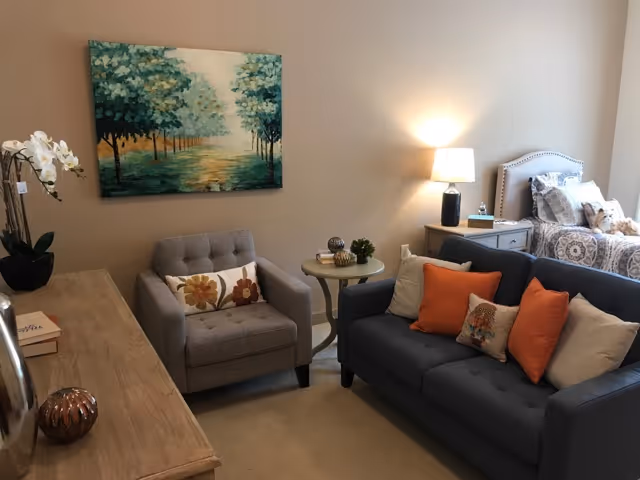 A cozy living area with a gray armchair and a dark blue sofa adorned with orange and beige pillows. A round side table with decorative items sits between the armchair and sofa. A wooden dresser with a potted orchid and books is on the left. In the background, there is a bed with patterned bedding and a nightstand with a lamp and water bottle. A painting of a tree-lined path hangs on the beige wall.