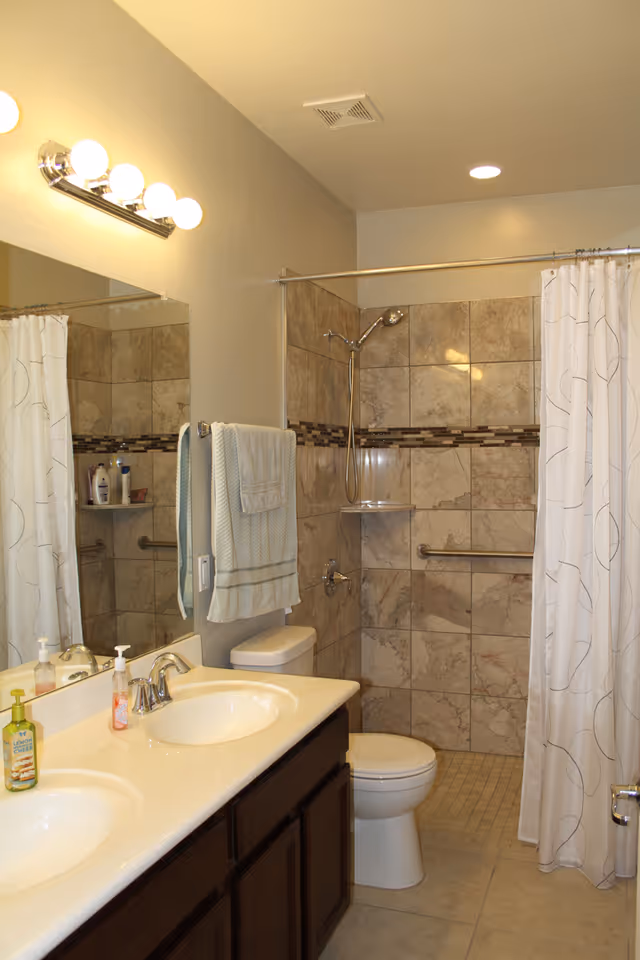 A clean and well-lit bathroom featuring a double sink vanity with two soap dispensers, a large mirror above, a towel hanging on the wall, a toilet, and a tiled walk-in shower with a shower curtain and grab bars.