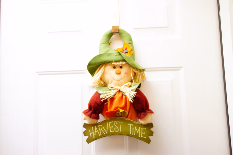 A decorative harvest scarecrow doll wearing a green hat hangs on a white door with a sign reading 'HARVEST TIME'.