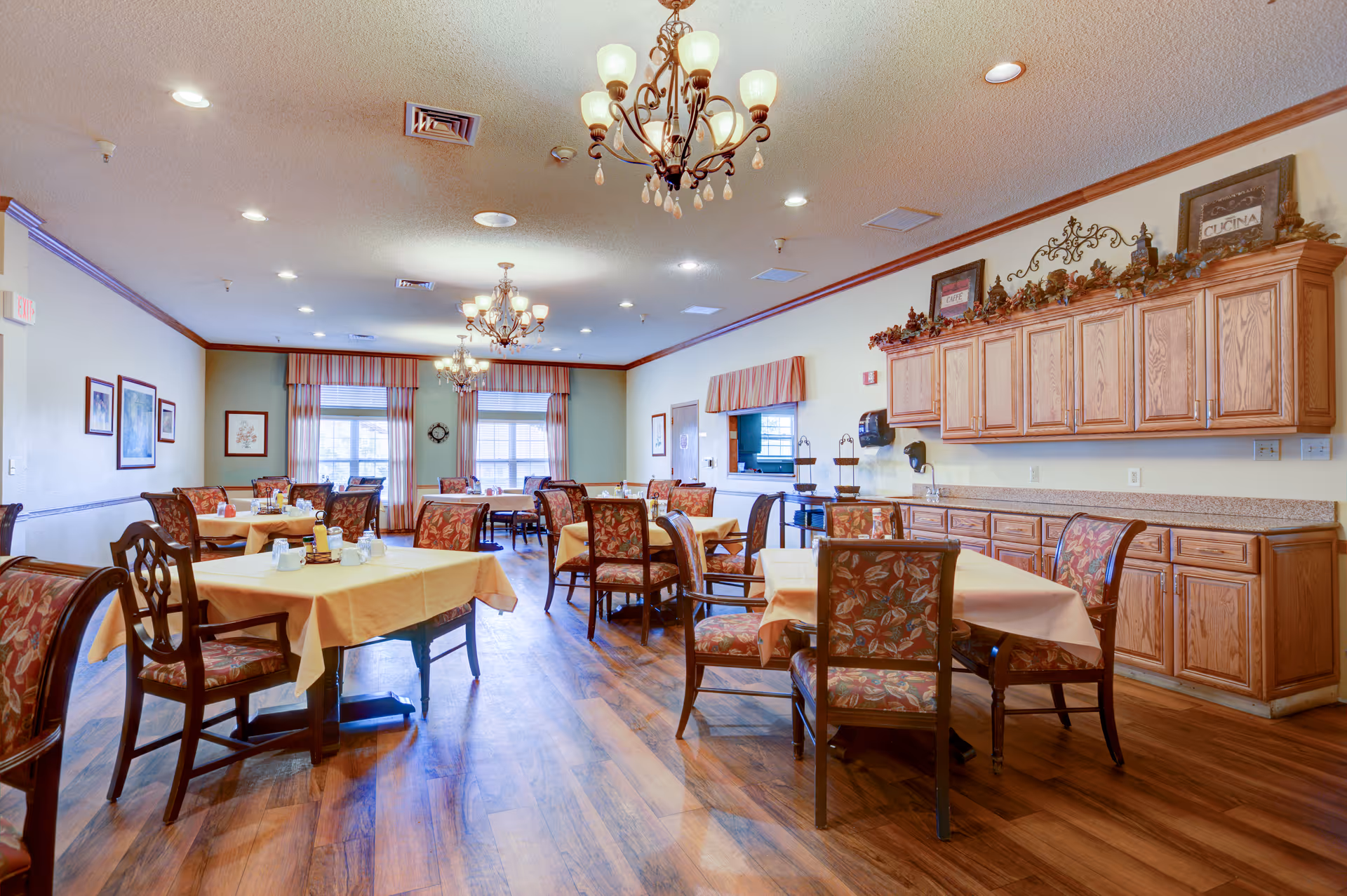 Bright dining room with multiple tables covered in yellow tablecloths, upholstered chairs, wooden cabinets, chandeliers, and windows.