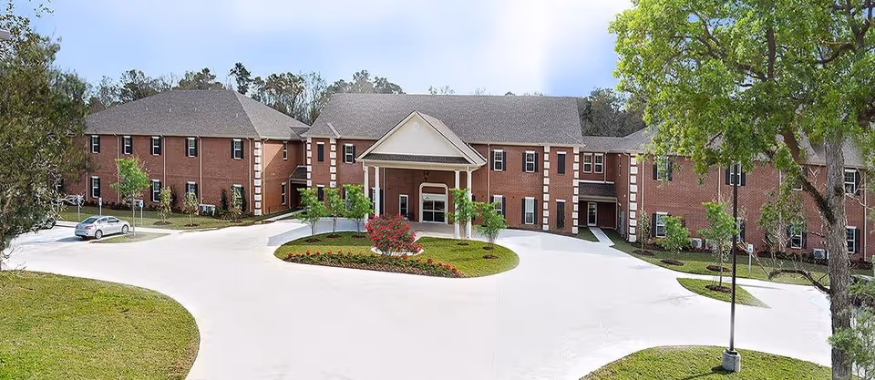 Front exterior view of a two-story brick building with a covered entrance and a circular driveway surrounded by landscaped greenery and trees under a clear sky.