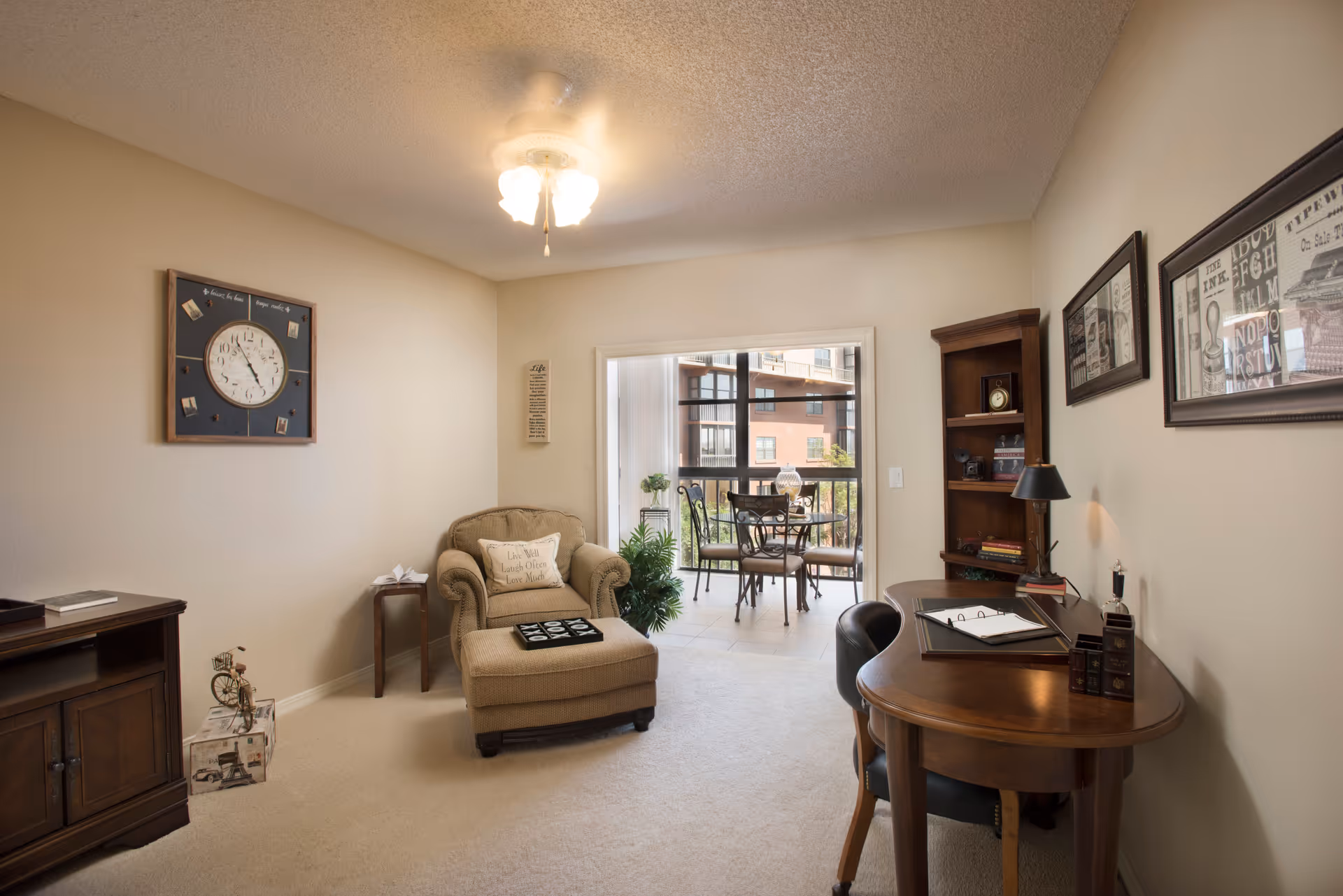 A cozy living room area with a beige armchair and ottoman, a wooden desk with a chair, and a wooden bookshelf. The room has light-colored walls and carpet, a ceiling fan with lights, and a large window leading to a balcony with a small table and chairs. The walls are decorated with framed pictures and a clock.