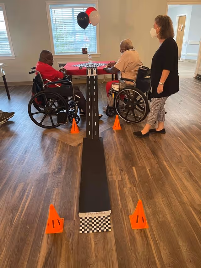 Two elderly men in wheelchairs sitting at a table with a red tablecloth and balloons in the background. A woman wearing a face mask stands nearby. In front of them is a small race track setup on the floor with orange cones marking the area and a checkered finish line.