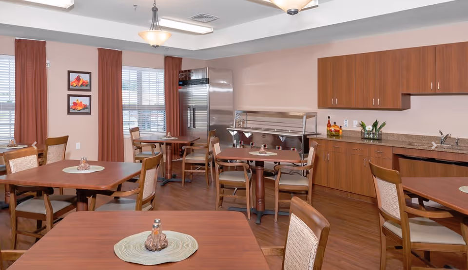 A dining area in a senior living facility with several wooden tables and chairs arranged neatly. The room has large windows with brown curtains, framed artwork on the wall, and a kitchen area with wooden cabinets, a countertop, a sink, and a stainless steel refrigerator. There is also a buffet serving station along one wall.