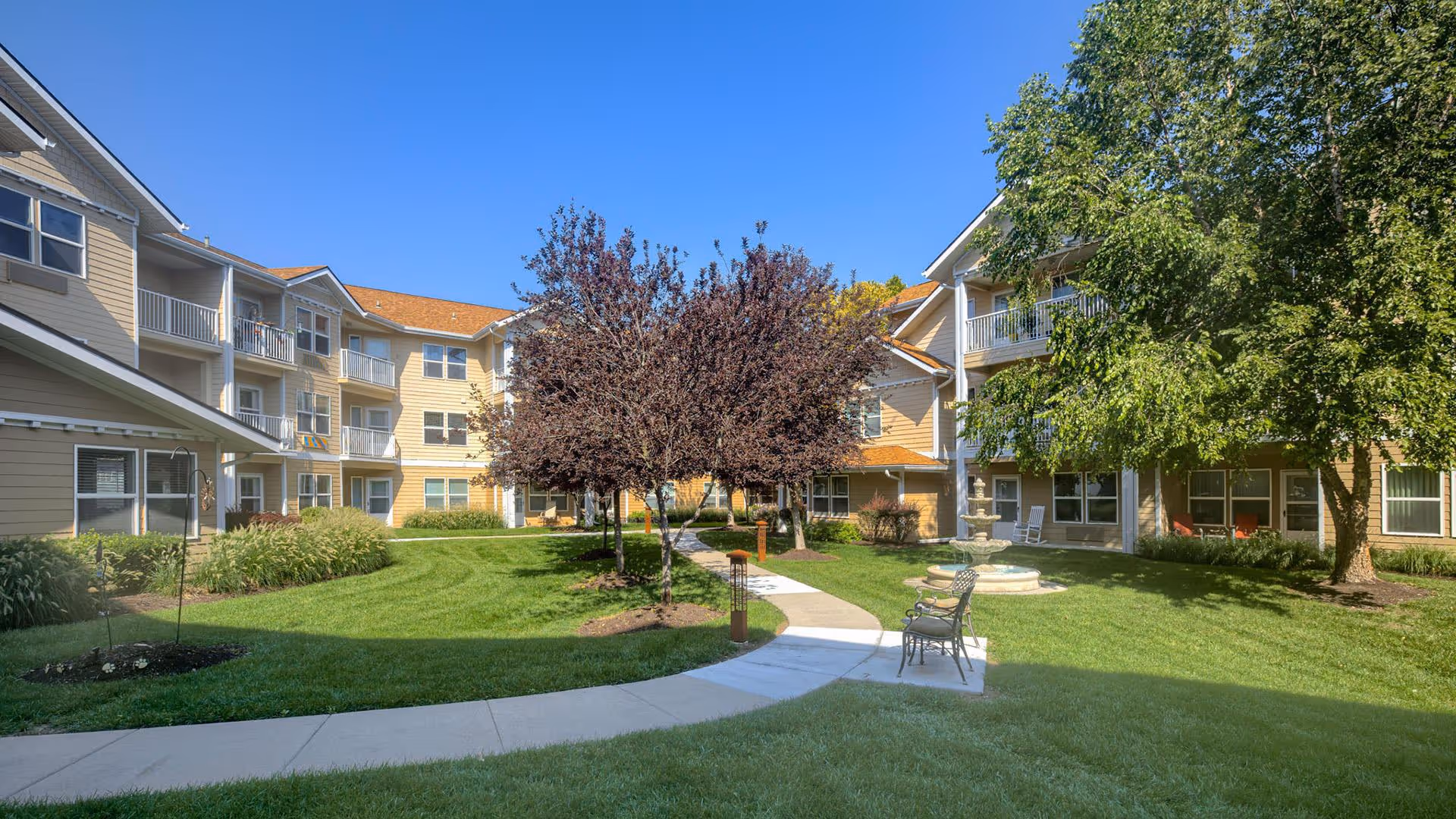 Outdoor courtyard area of Holiday Rolling Hills Ranch featuring a well-maintained lawn, paved walkways, several trees including some with purple leaves, a water fountain, and benches. The courtyard is surrounded by a multi-story residential building with balconies and windows under a clear blue sky.