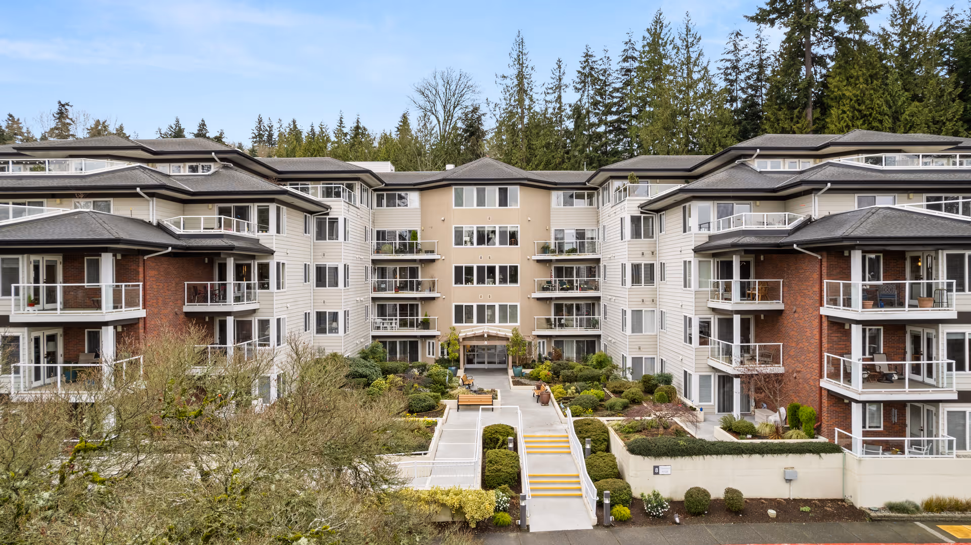 Exterior view of a multi-story senior living facility building with balconies, surrounded by landscaped greenery and trees in the background under a clear sky.
