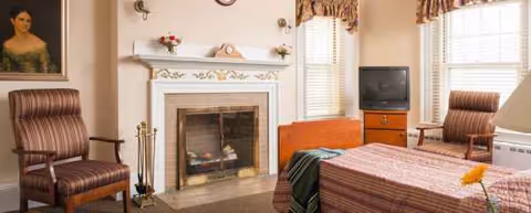Cozy nursing home bedroom featuring a bed, two chairs, a fireplace, and a TV.