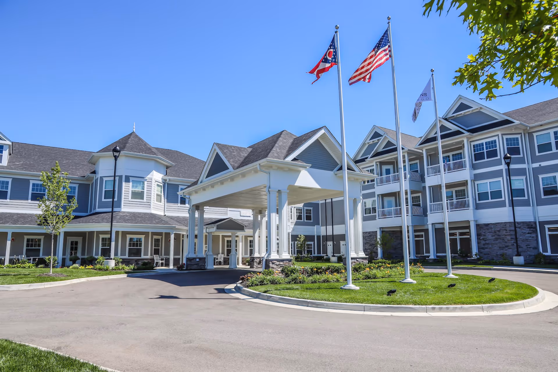Exterior view of Traditions at Camargo senior living facility with a covered entrance, three flagpoles displaying the Ohio state flag, the American flag, and a facility flag, surrounded by well-maintained landscaping and a clear blue sky.