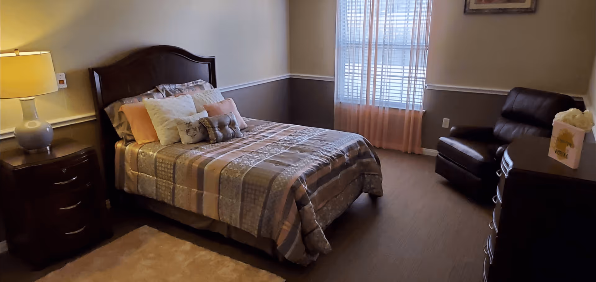 A cozy bedroom with a neatly made bed featuring a patterned quilt and multiple pillows. To the left of the bed is a wooden nightstand with a lamp. A window with sheer pink curtains lets in natural light. On the right side of the room, there is a dark leather armchair and a wooden dresser with a decorative item on top.