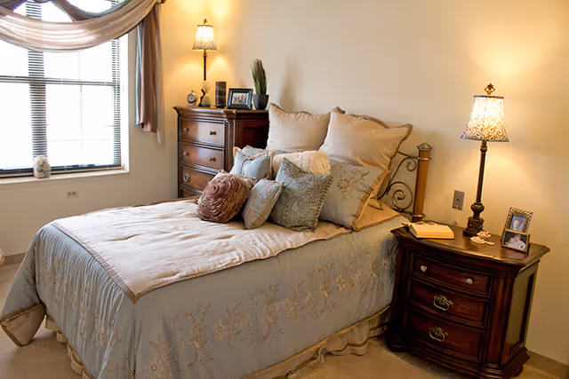 A cozy bedroom with a neatly made bed featuring multiple decorative pillows and a light blue bedspread with floral embroidery. There is a wooden nightstand with a lamp, a book, and a framed photo on the right side of the bed. On the left side, there is a wooden dresser with a lamp, framed photos, and a small plant. A window with blinds and curtains is visible on the left wall.