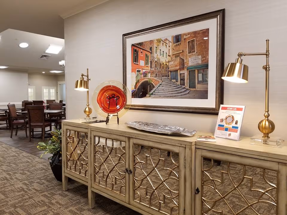 Interior view of a senior living facility showing a decorative sideboard with two gold lamps, a red and gold decorative plate, a silver tray, and a framed picture of a European street scene above it. In the background, there are tables and chairs arranged in a dining area.