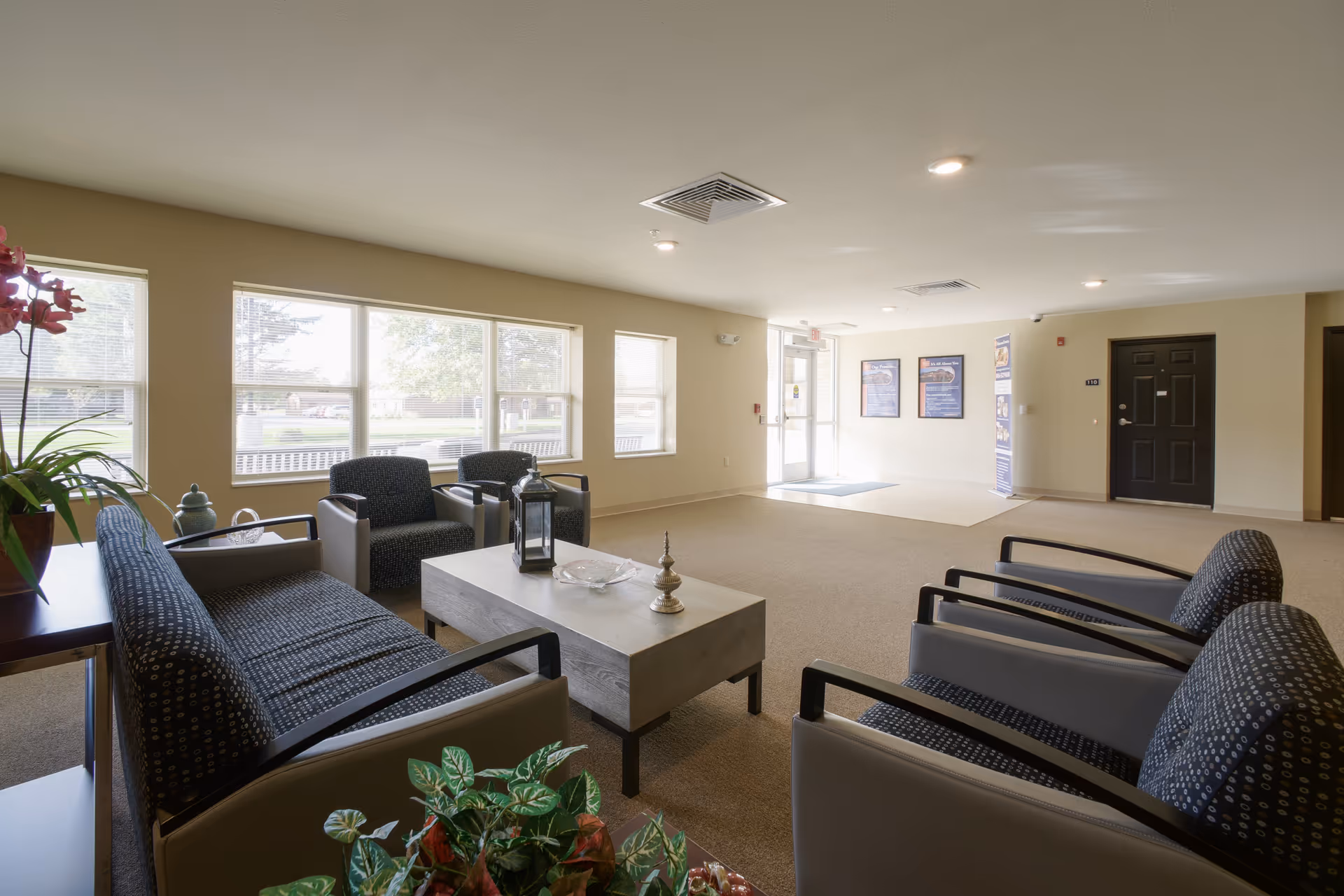 A bright and spacious senior living facility lounge area with multiple armchairs and a sofa arranged around a rectangular coffee table. Large windows on one side let in natural light, and there are plants and decorative items on the table and side surfaces. The entrance door and informational posters are visible in the background.