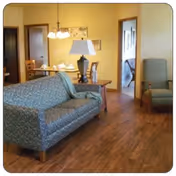 A cozy living room area with a patterned blue sofa, a wooden side table with a lamp, and a green armchair. In the background, there is a dining table with chairs and a hanging light fixture. Doorways lead to other rooms, including a bedroom.