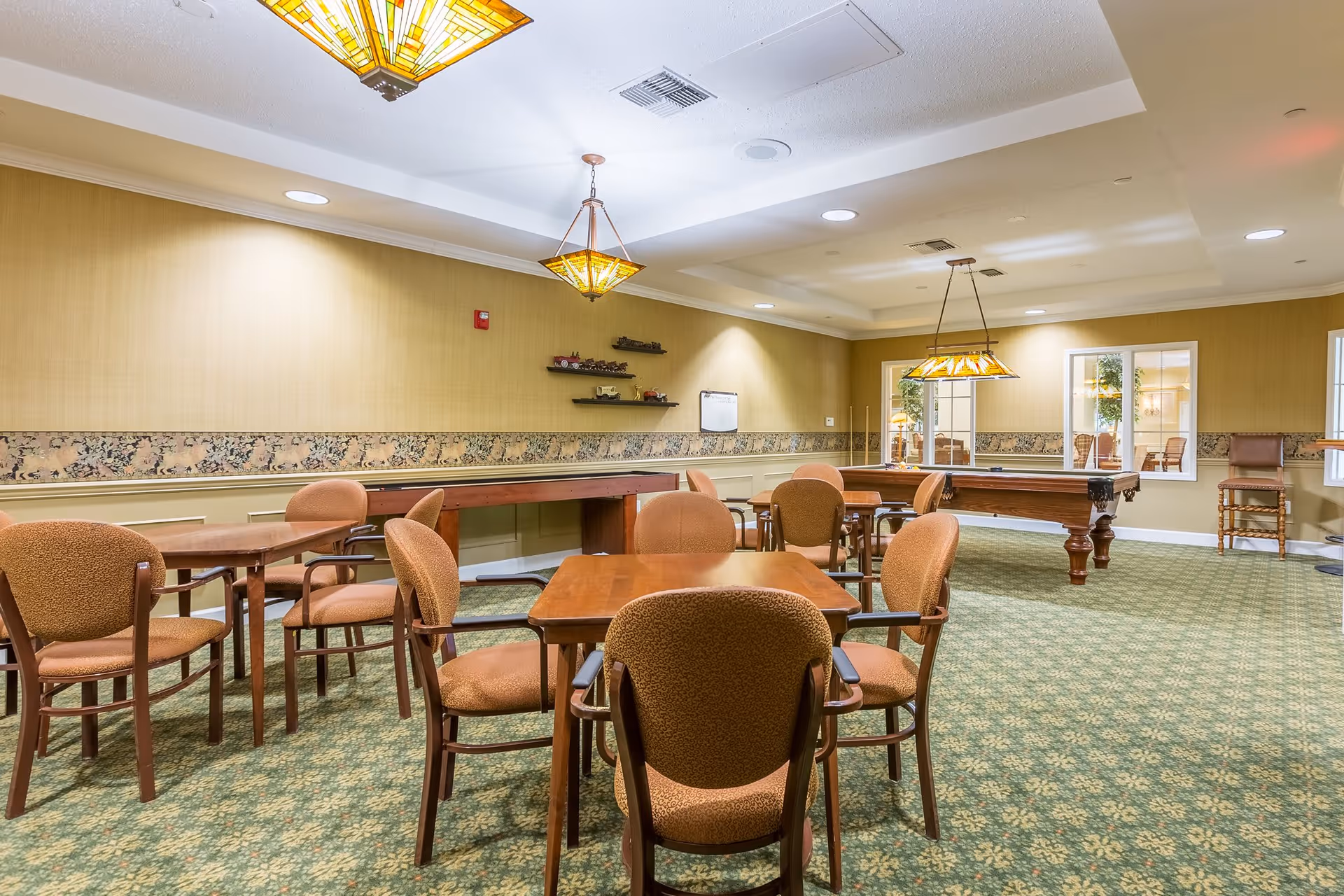 A spacious recreational room with multiple tables and chairs arranged for seating. The room features a pool table with hanging stained glass lights above it, patterned green carpet, beige walls with a floral border, and shelves with decorative items. There are windows looking into another room and additional chairs along the walls.
