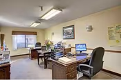 An office room with two desks, office chairs, a computer, paperwork, and a window with a view outside. The walls are light-colored with a framed picture and some notices posted. The room is lit by ceiling lights and wall sconces.