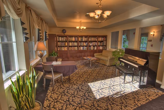 A cozy living room area in Chateau Valley Center Retirement featuring a patterned carpet, a piano with a bench, a beige sofa, a coffee table, a bookshelf filled with books, a table with a chess set, a lamp, and large windows with curtains letting in natural light.