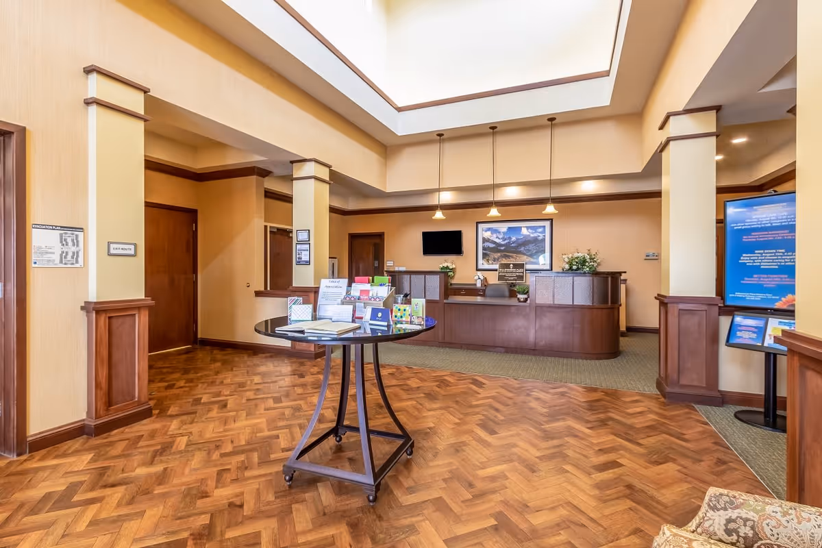 Reception area of Willowbrook Place featuring a wooden front desk with a computer and flowers, a round table in the center with brochures and books, warm beige walls, wooden flooring with a herringbone pattern, and a high ceiling with recessed lighting and hanging pendant lights.