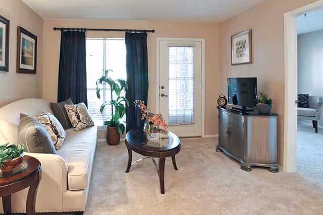 Cozy living room with a beige sofa, oval glass-top coffee table, TV on a wooden cabinet, and a window and door with dark curtains.