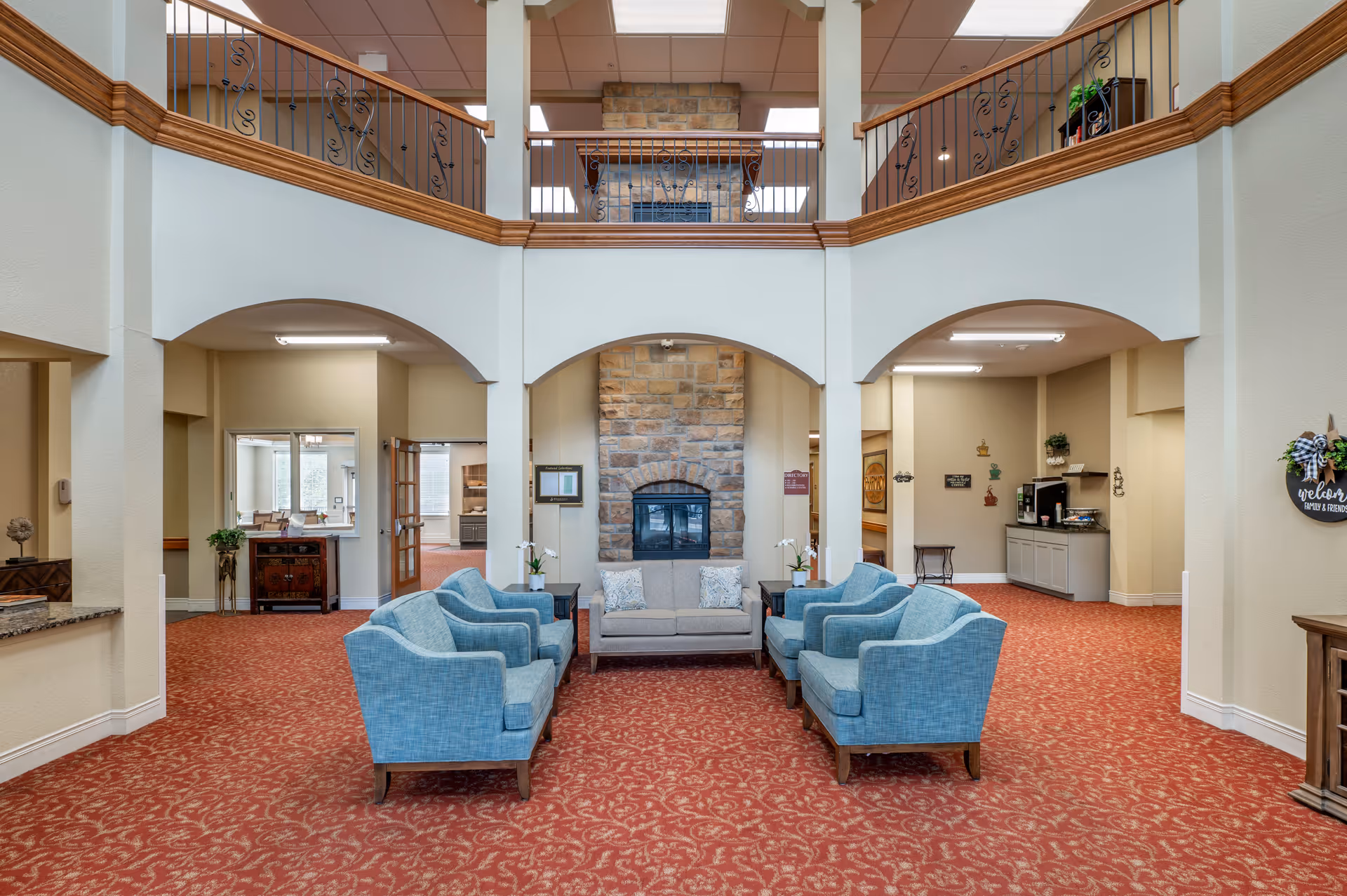 Two-story communal lounge with blue armchairs and a central sofa facing a stone fireplace beneath an open upper balcony.