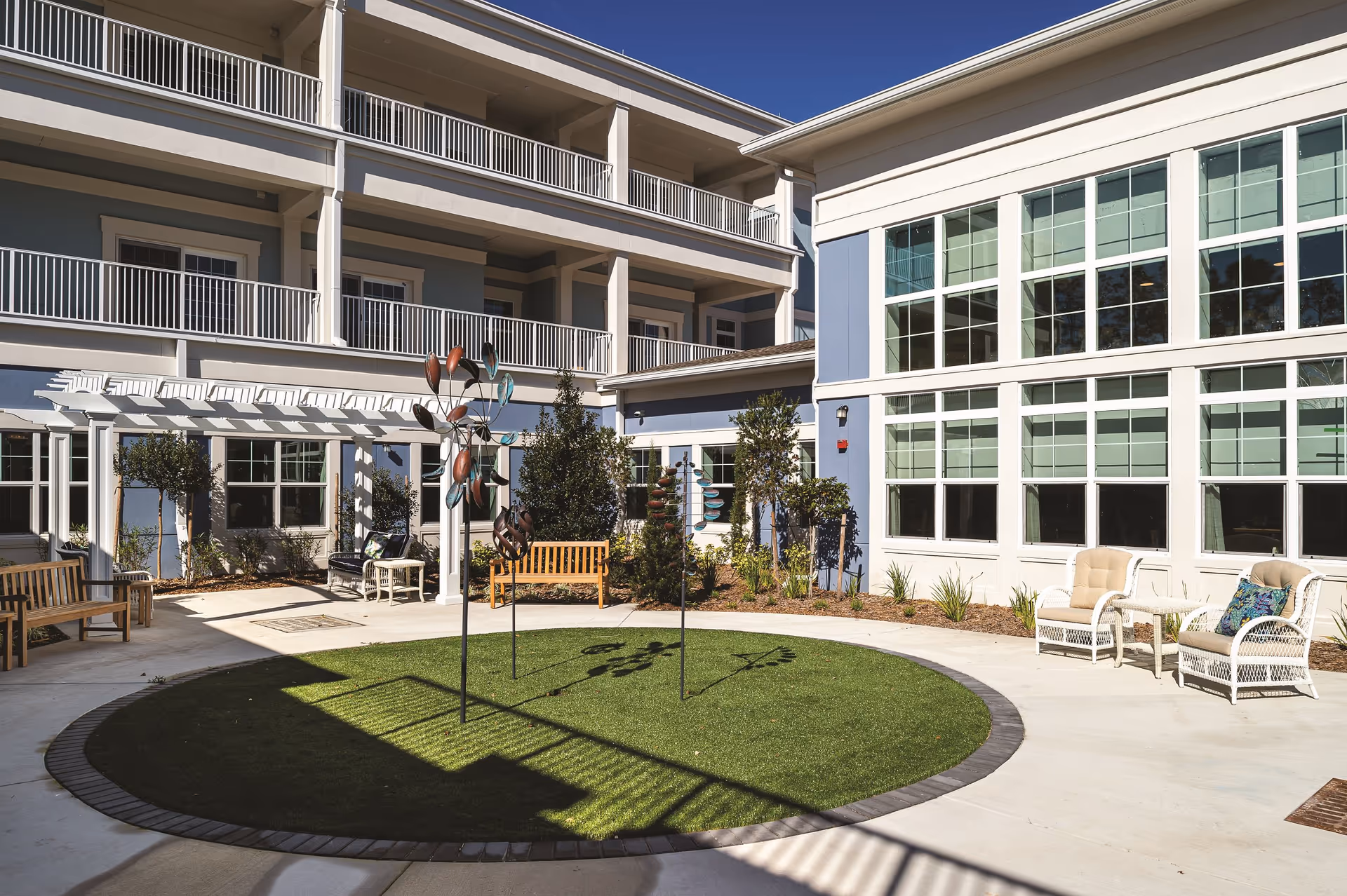 Outdoor courtyard area at Sancerre at Palm Coast featuring a circular patch of artificial grass with decorative metal sculptures, surrounded by a concrete walkway. There are wooden benches, white wicker chairs with cushions, and a white pergola attached to the building. The building has multiple floors with balconies and large windows, under a clear blue sky.