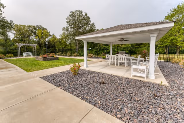 Outdoor patio area with a white covered pavilion featuring a ceiling fan and white outdoor furniture including a table and chairs. Surrounding the pavilion is a landscaped area with rocks and a concrete walkway. In the background, there is a green lawn with a white bench under a wooden pergola and trees.