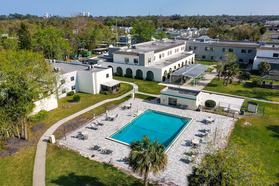 Aerial view of a senior living facility featuring a rectangular outdoor swimming pool surrounded by lounge chairs and tables with umbrellas. The pool area is paved and enclosed by a low fence. Surrounding the pool are green lawns, palm trees, and several white buildings with flat roofs and arched walkways. The background shows more trees and distant buildings under a clear sky.