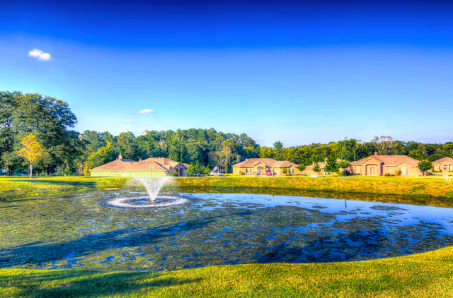 A scenic outdoor view of a pond with a water fountain in the center, surrounded by green grass and trees. In the background, there are several single-story buildings under a clear blue sky.