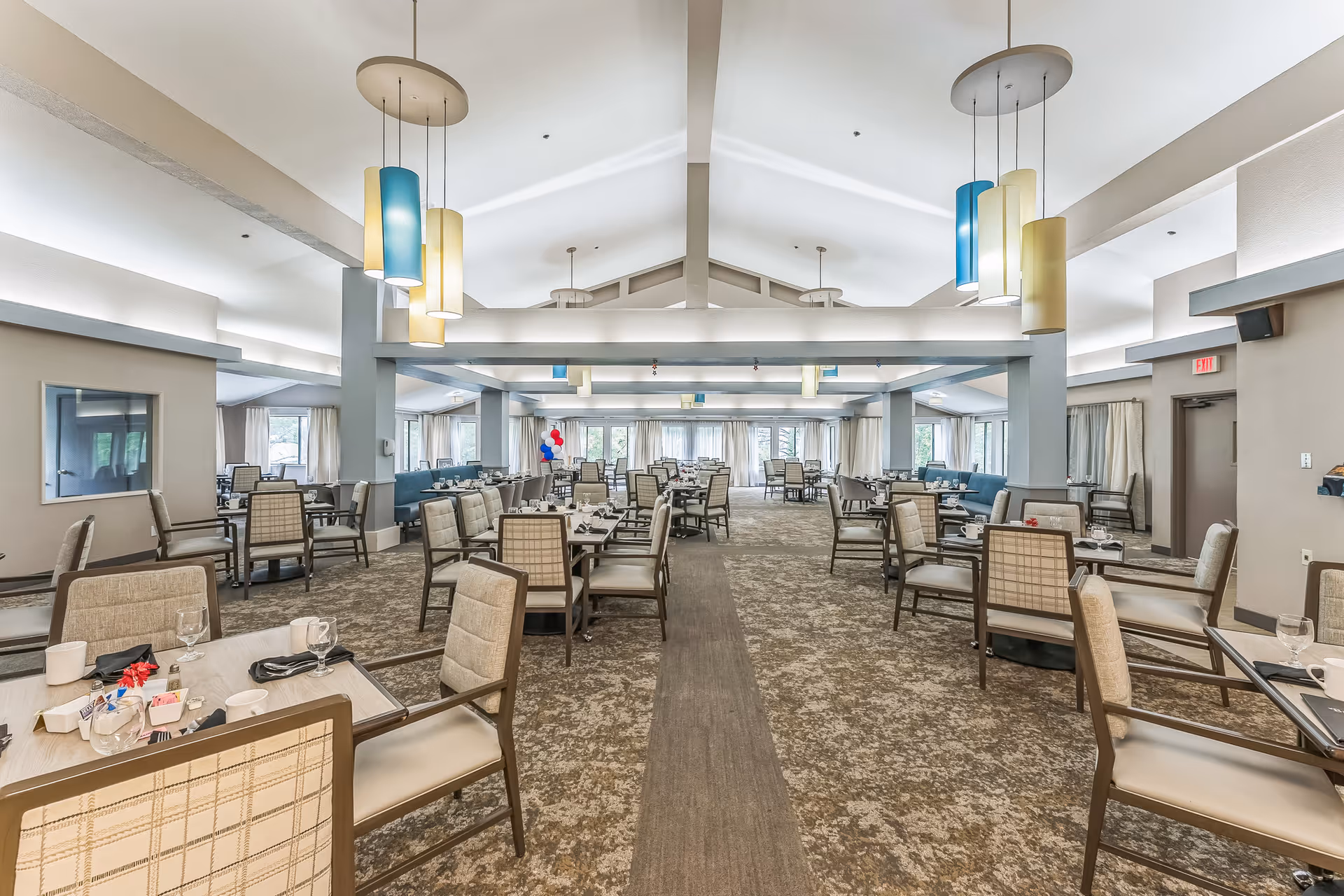 A spacious dining room with multiple tables and chairs arranged neatly. The room features high vaulted ceilings with modern pendant lights in blue and yellow hues. Large windows with curtains allow natural light to fill the space. The carpeted floor has a patterned design, and the tables are set with glassware and napkins.