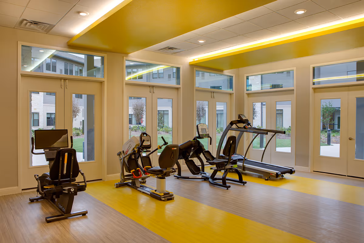 A bright fitness room with large windows and glass doors showing an outdoor courtyard. The room contains exercise equipment including a recumbent bike, a rowing machine, a leg press machine, and a treadmill. The floor has a yellow and wood pattern, and the ceiling features recessed lighting with a yellow accent.
