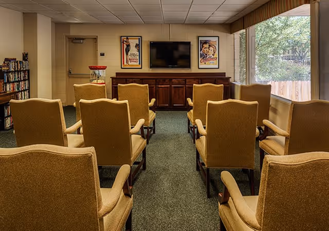 A small media room with rows of upholstered armchairs facing a wall-mounted TV and wooden cabinet, framed posters on the wall and a large window to the right.