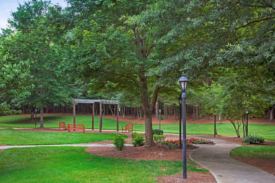 Park-like outdoor courtyard with a winding paved path, benches under a pergola, lampposts, and mature trees.