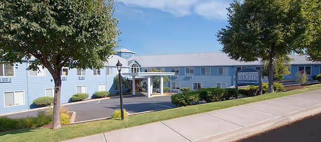 Exterior view of a two-story senior living facility building with light blue siding, white trim, and a covered entrance. There are trees, shrubs, and a sidewalk in the foreground, along with a sign that reads 'Heights'.