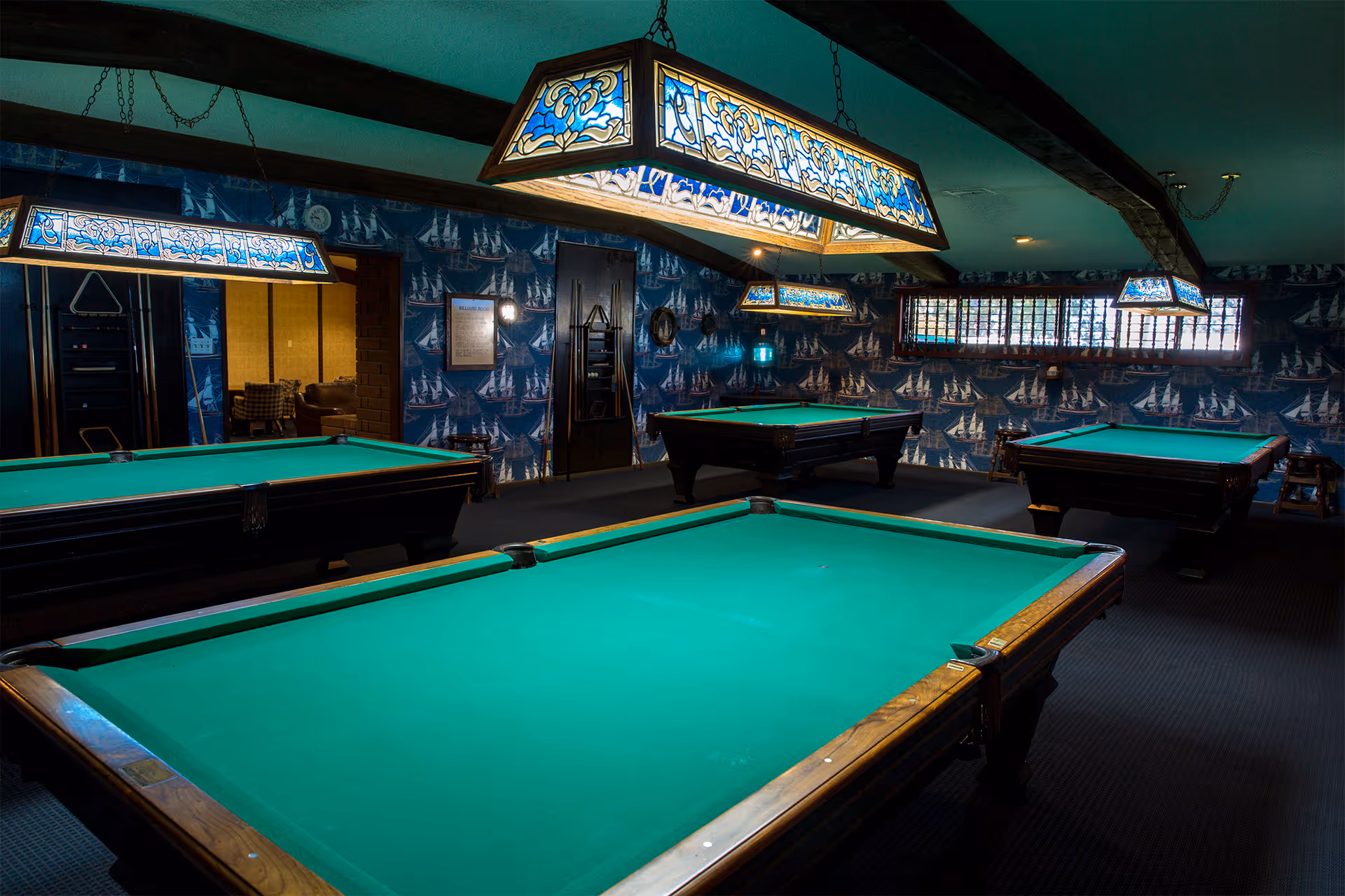 Interior view of a recreational room with four green pool tables under decorative stained glass hanging lights. The walls are covered with blue wallpaper featuring white sailboats, and there is a window with bars on the right side. A doorway leads to another room with seating visible.