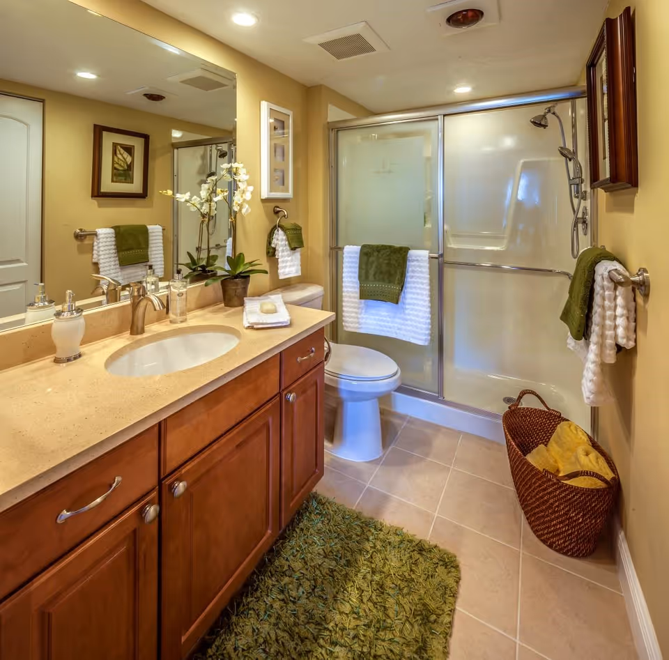A well-lit bathroom featuring a beige countertop with a sink, a large mirror, wooden cabinets, a toilet, and a glass-enclosed shower with sliding doors. The bathroom is decorated with green and white towels, a green rug, a potted orchid plant on the countertop, framed artwork on the walls, and a woven basket with yellow towels on the floor.