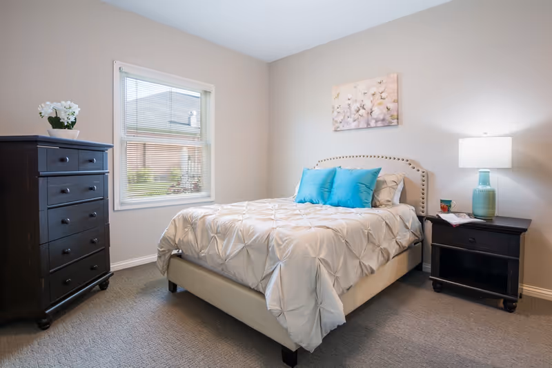 A neatly made bed with a beige tufted headboard and white comforter accented by three bright blue pillows in a bedroom. To the right of the bed is a dark wooden nightstand with a turquoise lamp, a mug, and a book. To the left is a matching dark wooden dresser with a white flower pot on top. A window with blinds shows an exterior view of a neighboring building. A floral painting hangs on the wall above the bed.