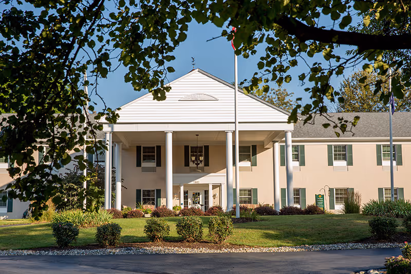 Front exterior view of a two-story senior living facility building with white columns and green shutters, surrounded by landscaped bushes and trees under a clear blue sky.