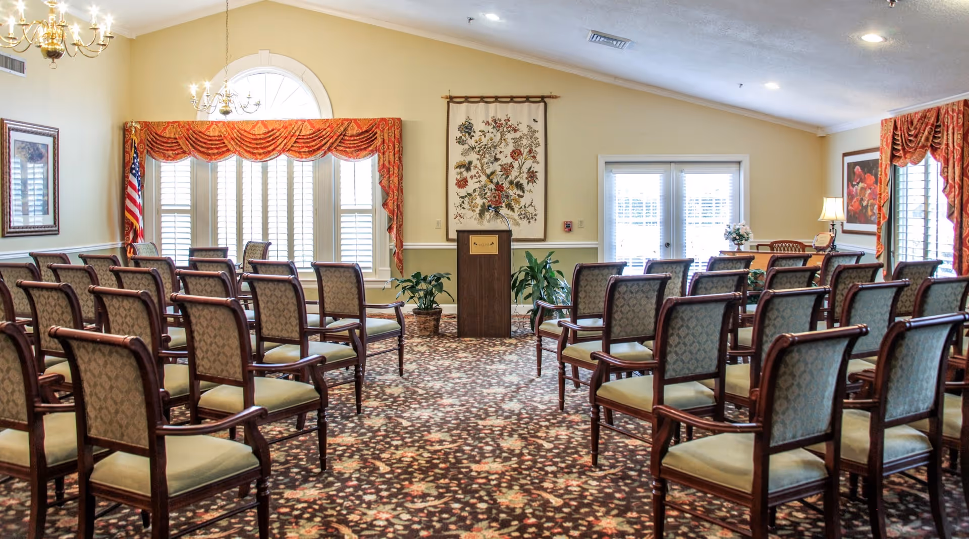 A well-lit meeting or event room with rows of upholstered chairs arranged facing a wooden podium. The room features patterned carpet, large windows with white shutters and red valances, an American flag, potted plants, framed artwork, and a table with a lamp and floral arrangement near the back.