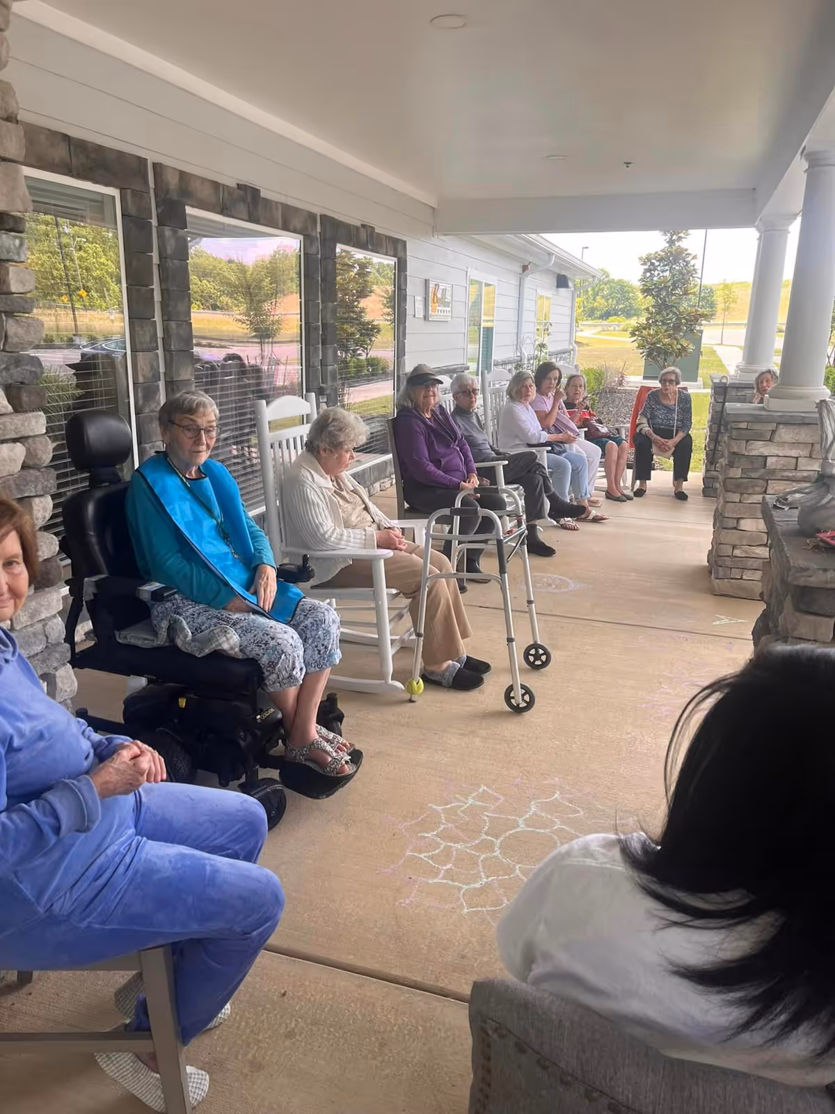 A group of elderly people sitting on rocking chairs and regular chairs on a covered porch outside a building. Some individuals use mobility aids like a walker and a wheelchair. The porch has stone pillars and windows along the wall, with chalk drawings on the concrete floor.