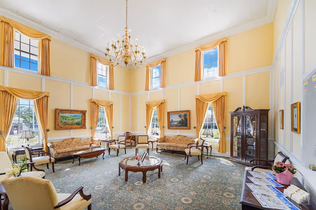 A spacious and elegant living room with high ceilings and large windows draped with yellow curtains. The room is furnished with vintage-style sofas, armchairs, and wooden coffee tables arranged on a patterned carpet. A chandelier hangs from the ceiling, and paintings adorn the walls. A glass display cabinet and a side table with brochures and a potted plant are also visible.