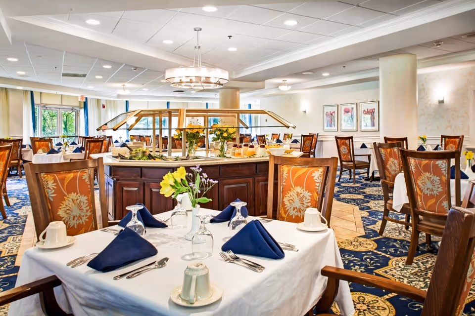 A spacious dining room in Bedford Court with tables covered in white tablecloths, each set with navy blue napkins, glassware, and silverware. The room features patterned carpet, wooden chairs with floral upholstery, and a central buffet station with flowers and food items. Large windows allow natural light to brighten the space.
