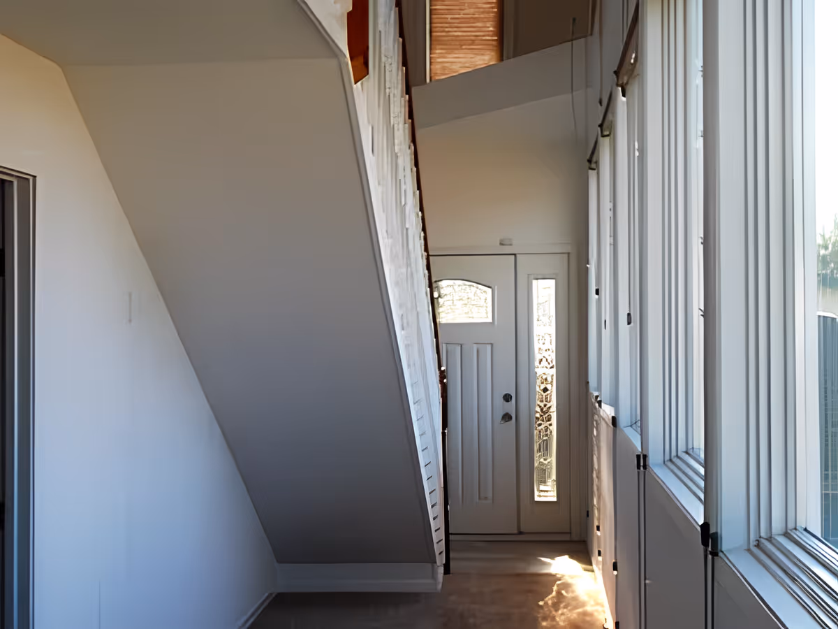 Bright narrow entry hallway showing the underside of a staircase, a front door with decorative glass, and tall windows along the right side.