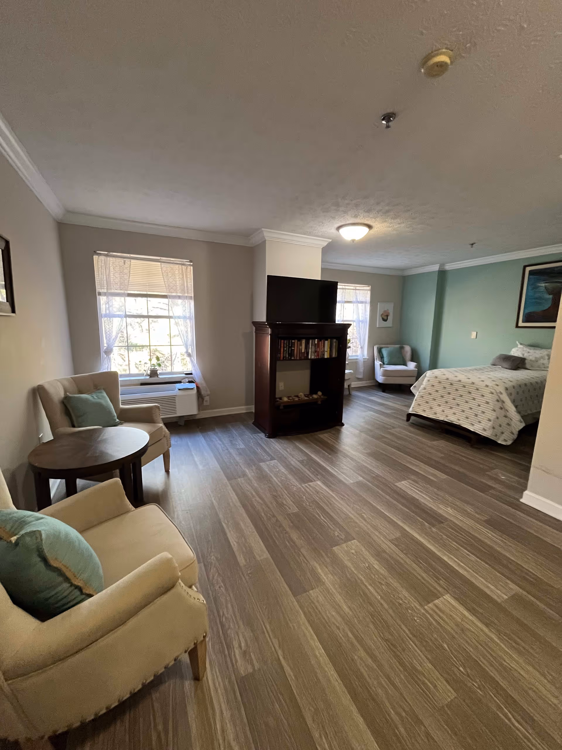 A spacious room with wood flooring featuring a sitting area with two beige armchairs and a round wooden table, a TV on a dark wooden stand with shelves holding books, and a bed with patterned bedding against a green accent wall. Two windows with sheer curtains allow natural light into the room.