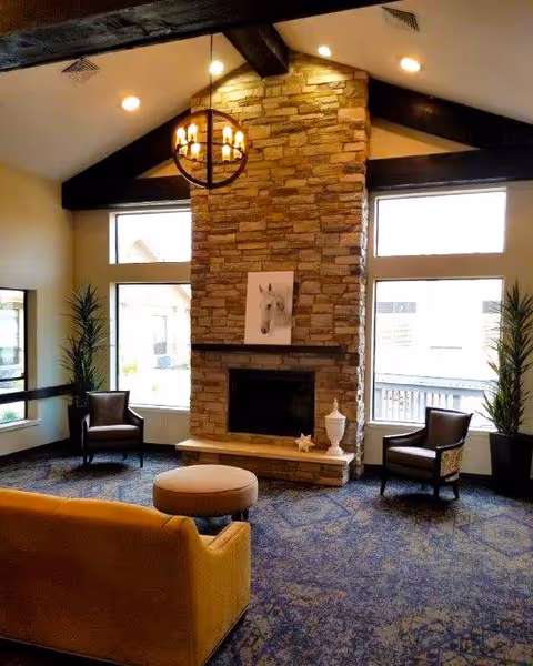 A cozy living room area with a high vaulted ceiling featuring dark wooden beams. The focal point is a tall stone fireplace with a painting of a horse above it. There are two large windows on either side of the fireplace letting in natural light. The room is furnished with a mustard yellow sofa, two dark armchairs, a round ottoman, and two tall potted plants near the windows. A modern chandelier hangs from the ceiling.