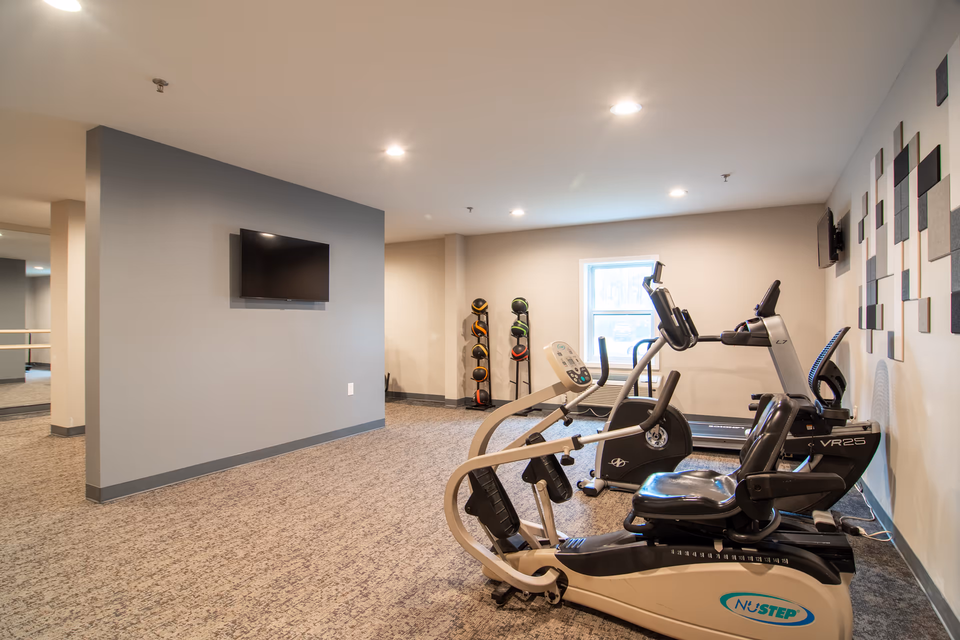 A fitness room in a senior living facility with exercise equipment including a recumbent bike, treadmill, and a rack of medicine balls. The room has carpeted floors, neutral-colored walls, a wall-mounted TV, and ceiling lights providing bright illumination.