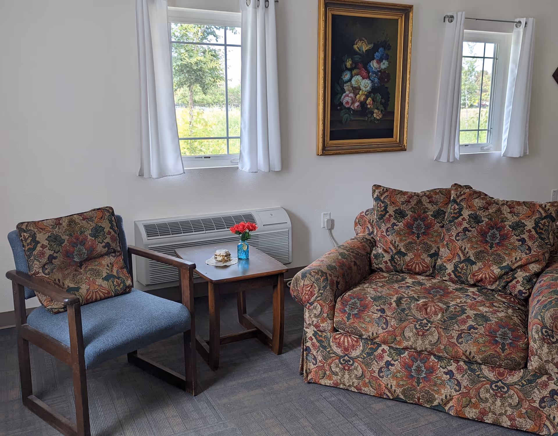 A cozy sitting area with a floral patterned armchair and a blue cushioned wooden chair with a matching floral pillow. Between them is a small wooden table holding a small vase with red flowers and a decorative item. Two windows with white curtains let in natural light, and a framed floral painting hangs on the wall above the armchair.